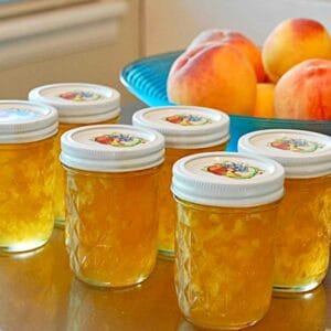 Eight jars of homemade peach preserves in front of a bowl of fresh peaches.