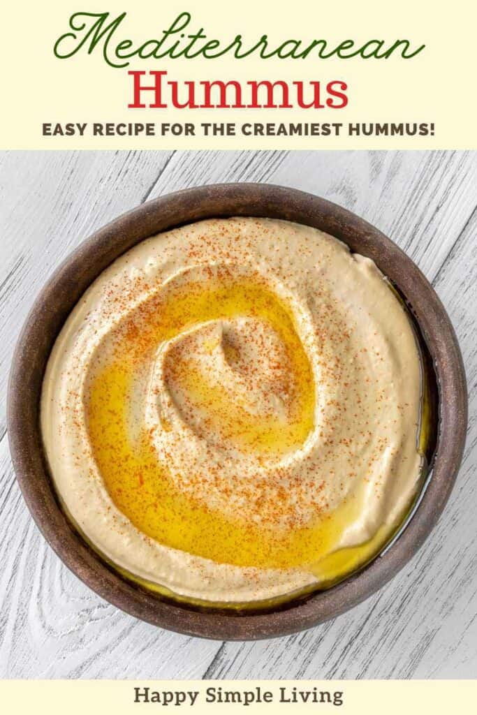 A bowl of creamy hummus drizzled with olive oil and sprinkled with paprika sits on a light wooden surface. Text above reads Mediterranean Hummus and Easy recipe for the creamiest hummus! Below: Happy Simple Living.