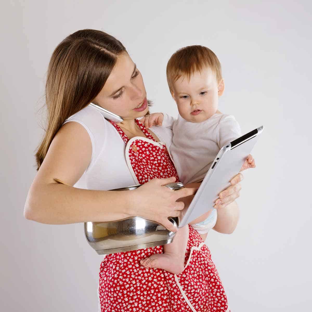 A woman holding a baby, a tablet, and a saucepan, trying to do too much.
