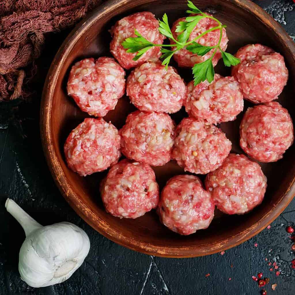 A batch of homemade uncooked meatballs in a wooden bowl, with a head of whole garlic nearby.
