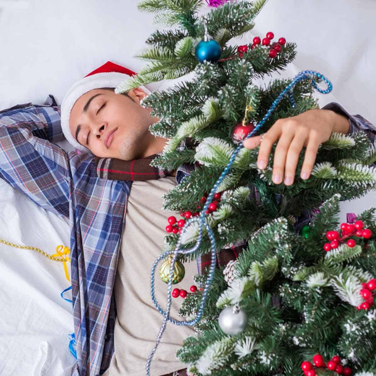 A person in a Santa cap sleeping next to a Christmas tree.
