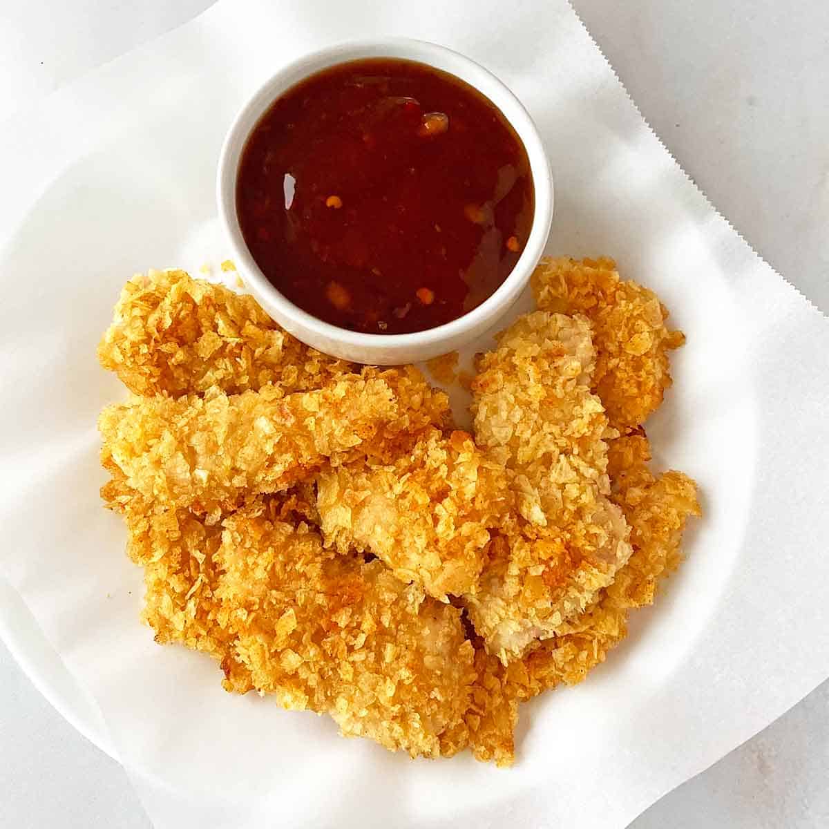A dozen pieces of golden brown potato chip chicken tenders on a plate with sweet chili sauce for dipping.