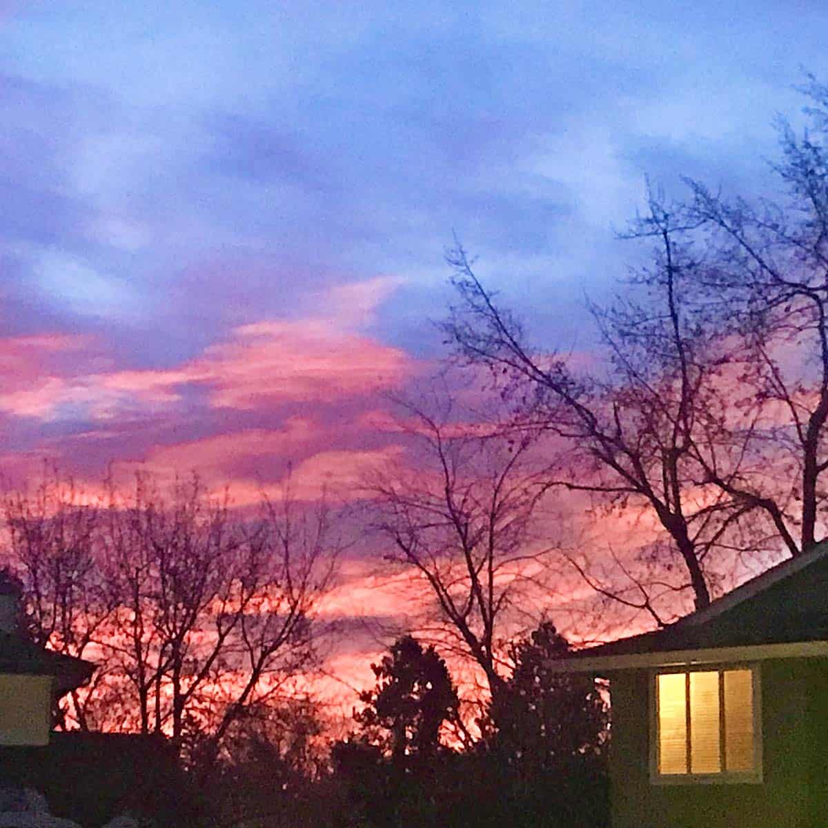 A beautiful sunrise with blue and pink clouds and a house to the right with a light on in the window.