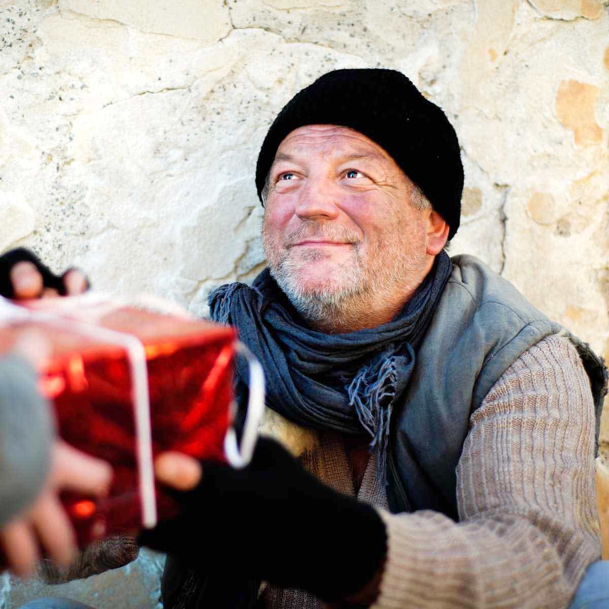 A homeless man smiling as he is handed a Christmas gift wrapped in red wrapping paper.