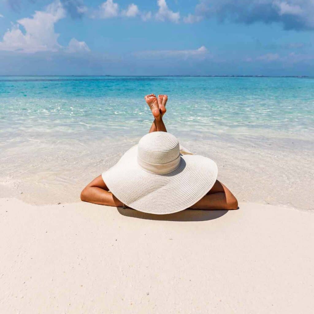 A woman wearing a giant sun hat and lying on the beach.