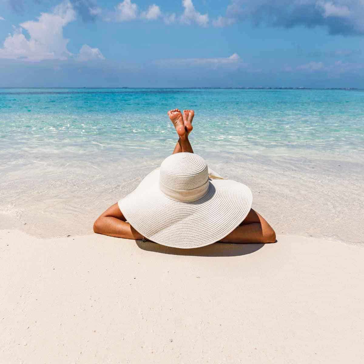 A woman wearing a large sun hat and lying on the beach. 