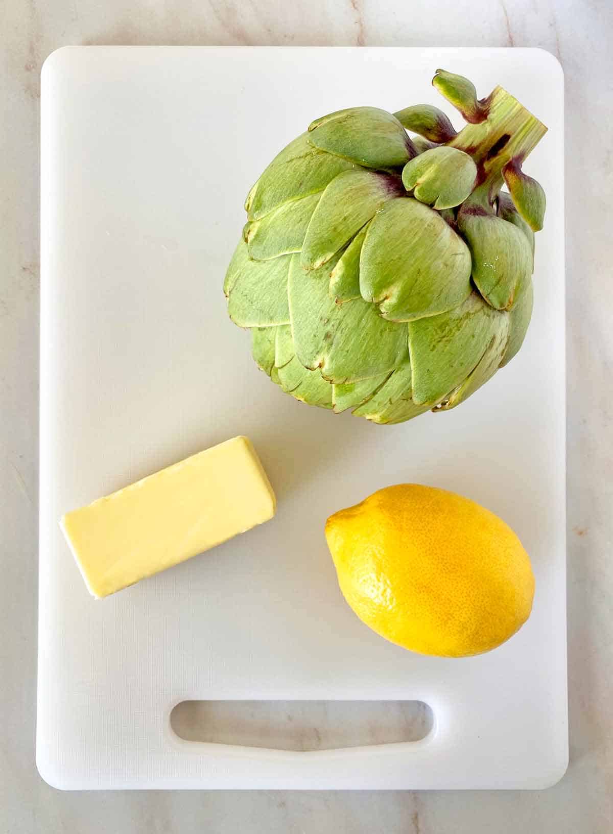 An artichoke, a stick of butter and a lemon on a cutting board.