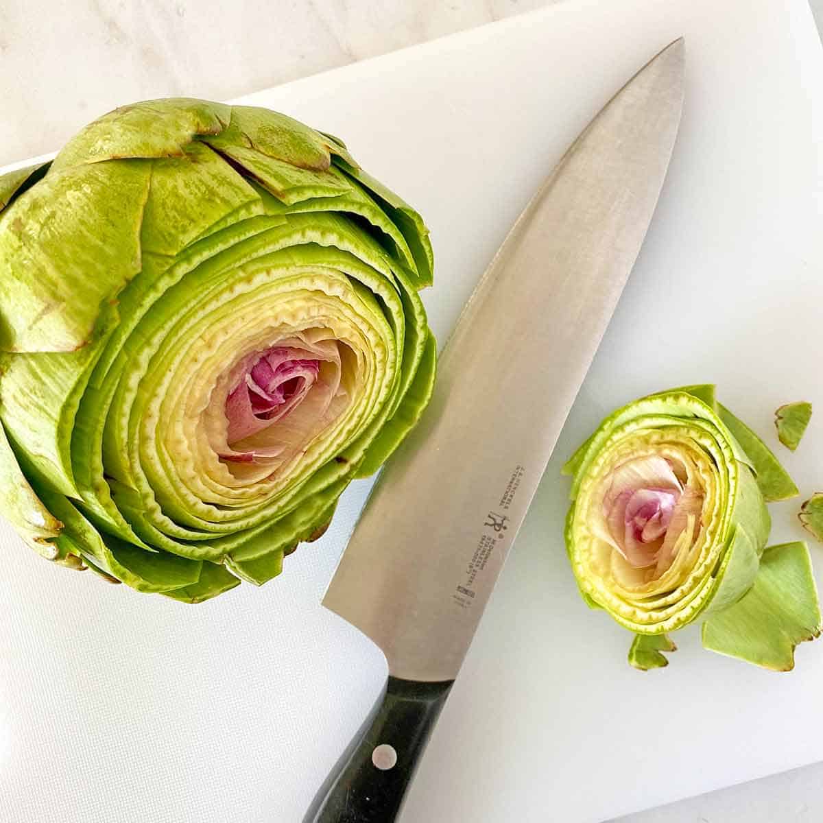 An artichoke with the top sliced off and a knife on a cutting board.