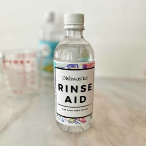 A bottle of homemade DIY dishwasher rinse aid, with a measuring cup and bottle of vinegar in the background.