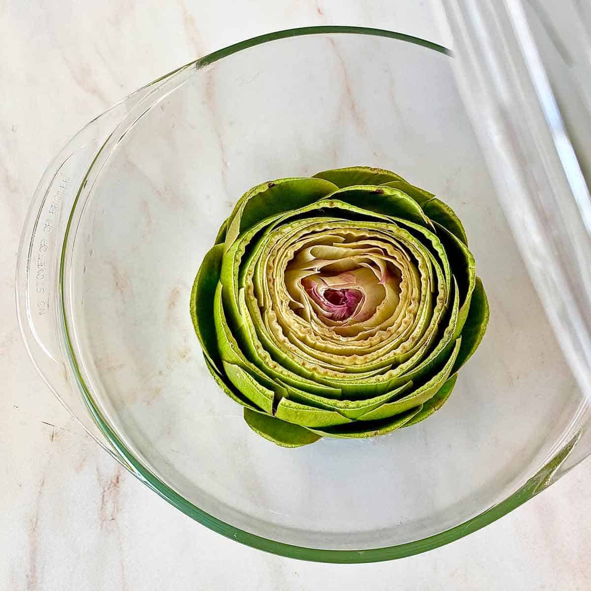 An artichoke in a microwave safe bowl ready for cooking.