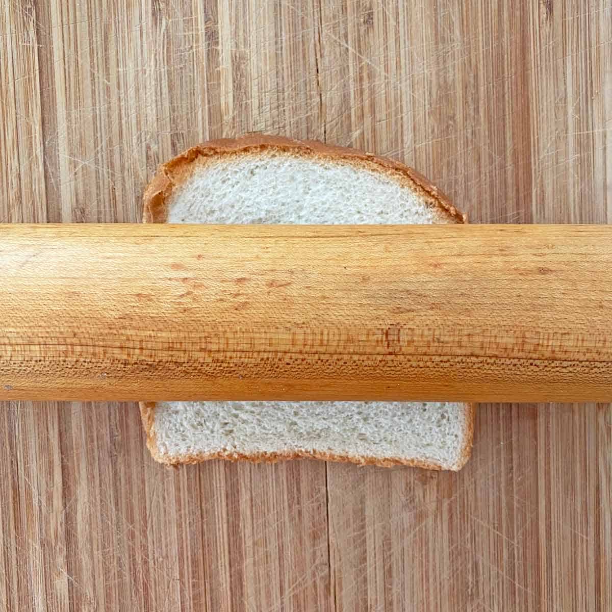 Rolling a piece of white bread with a rolling pin on a cutting board.