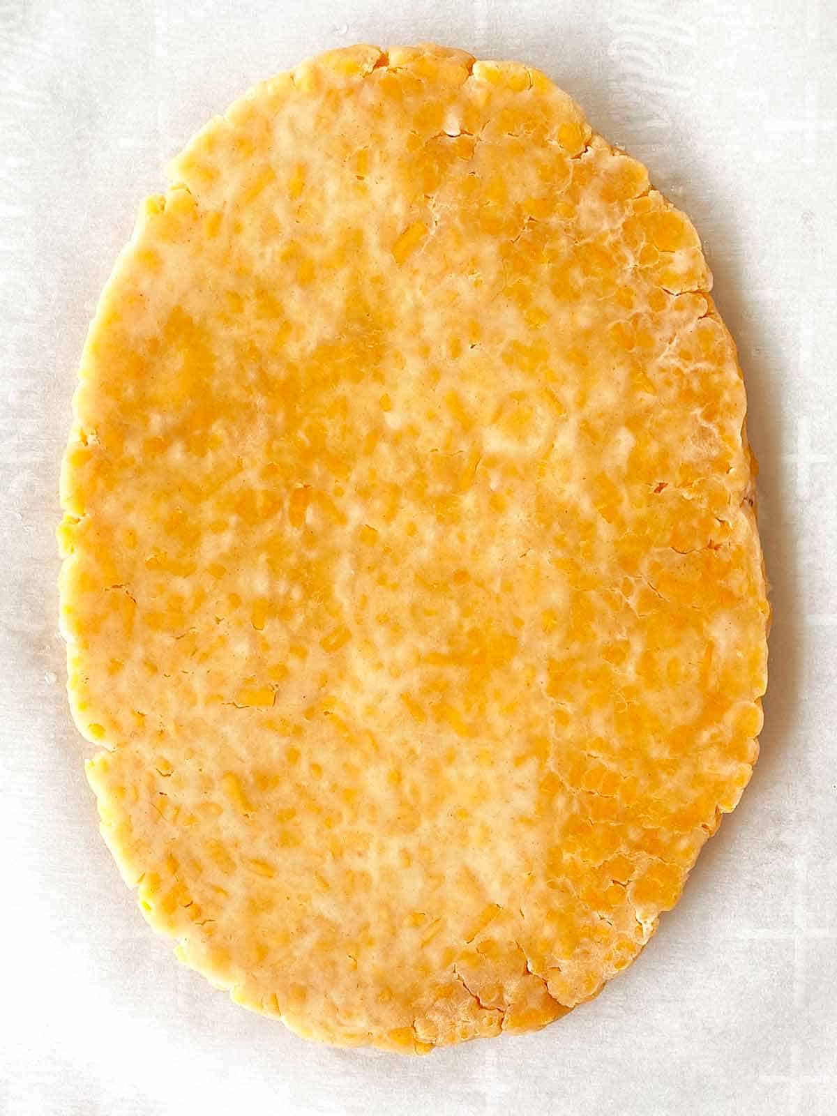 An oval of partially rolled cheez cracker dough on parchment paper.