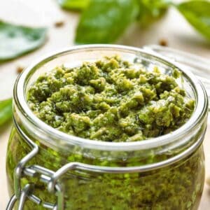 A jar of mint pesto sauce with basil leaves in the background.
