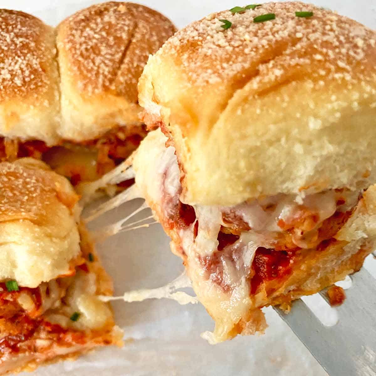 Lifting a meatball slider from the pan with melting cheese pulling apart.