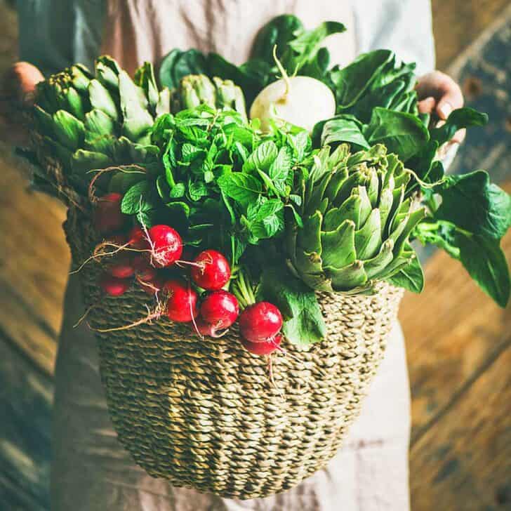 A woman holding a garden basket filled with radishes, onions, lettuce and artichokes.