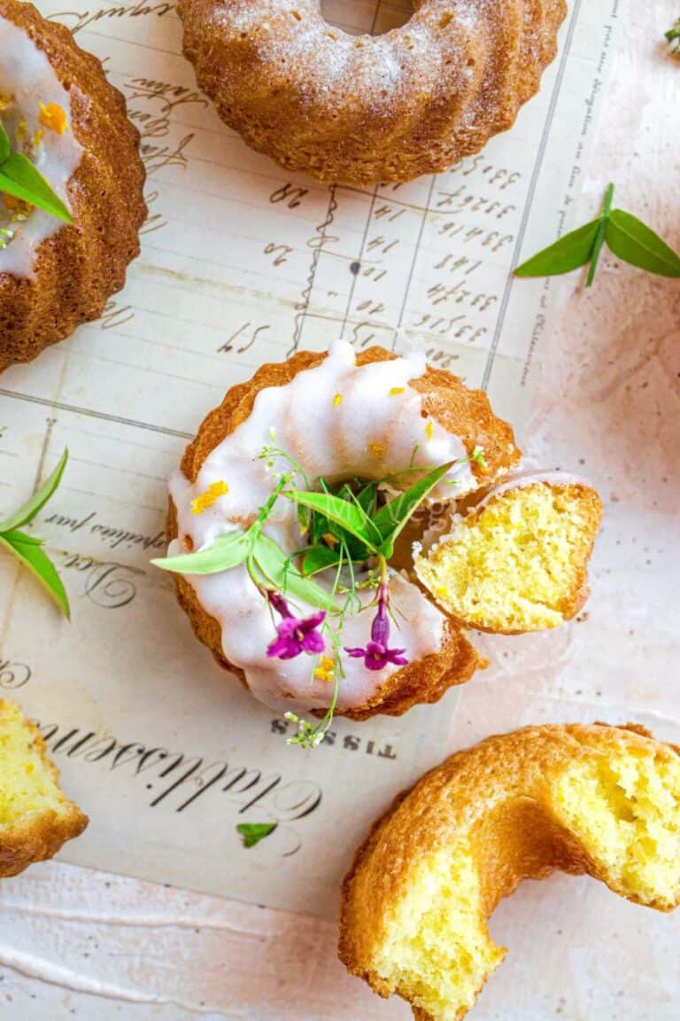 Mini bundt cakes on a vintage paper background. One is topped with white glaze and purple flowers, a slice cut out. A cut piece, a whole cake, and green leaves are scattered around. The surface is light pink.
