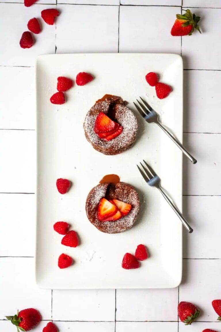 Two chocolate lava cakes topped with sliced strawberries and powdered sugar on a rectangular white plate. Two forks are placed beside the cakes. Fresh raspberries are scattered around on the plate and table.