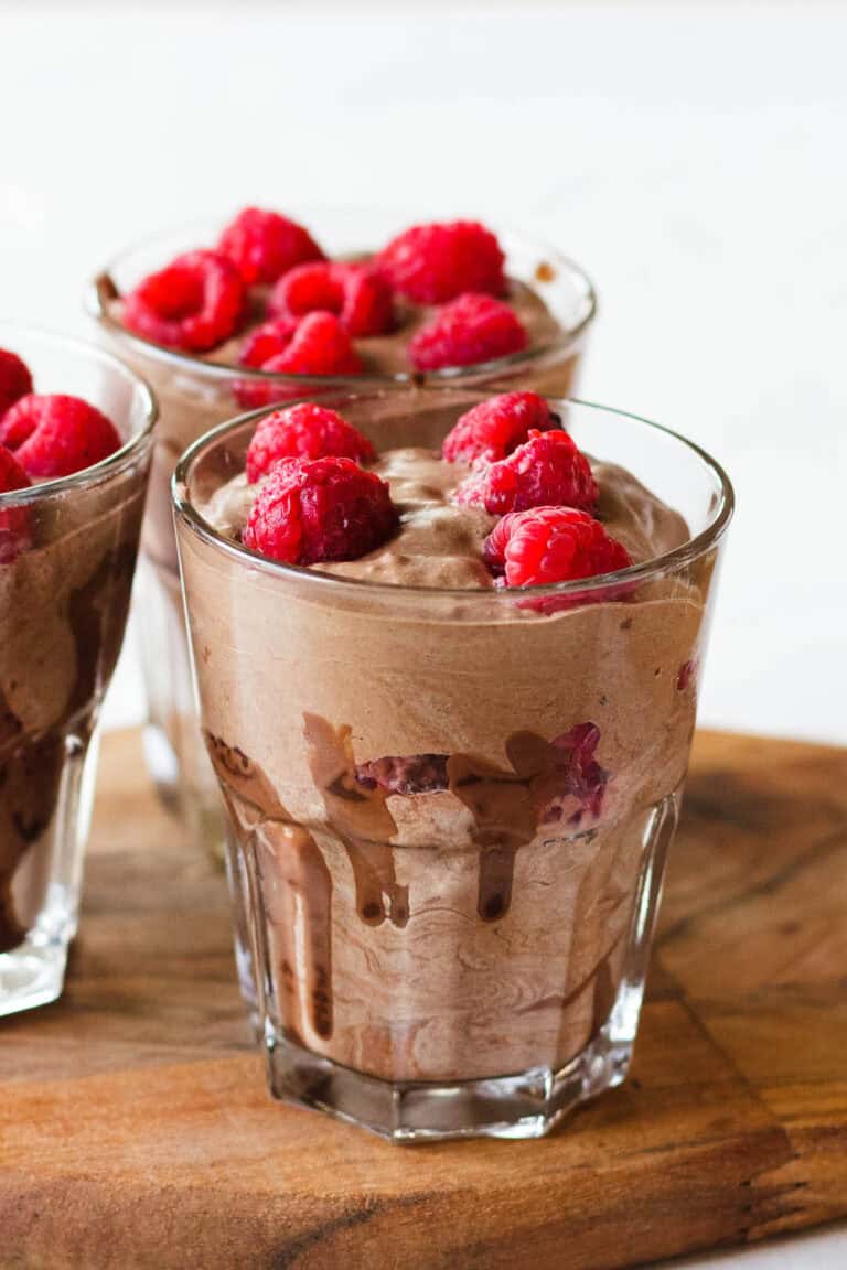 Three clear glasses filled with chocolate mousse topped with fresh raspberries. The mousse has a creamy, swirled texture and the raspberries add a vibrant red contrast. The glasses are placed on a wooden surface.