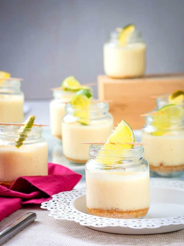 Small glass jars filled with creamy dessert topped with lime slices are arranged on a doily-covered plate. A red napkin and a spoon are nearby. A wooden block holds more jars in the background, creating a neat presentation.