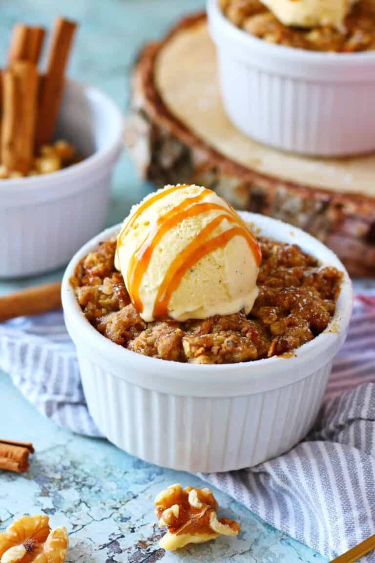 A ramekin filled with apple crisp topped with a scoop of vanilla ice cream drizzled with caramel sauce. Its placed on a striped napkin, with cinnamon sticks and walnuts scattered nearby on a textured blue surface.