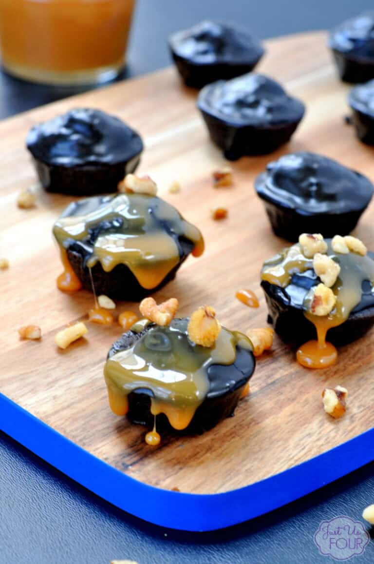 Mini chocolate caramel tarts topped with walnut pieces are arranged on a wooden board with a blue edge. A glass of caramel sauce is visible in the background. The tarts have a glossy chocolate surface with caramel dripping down the sides.