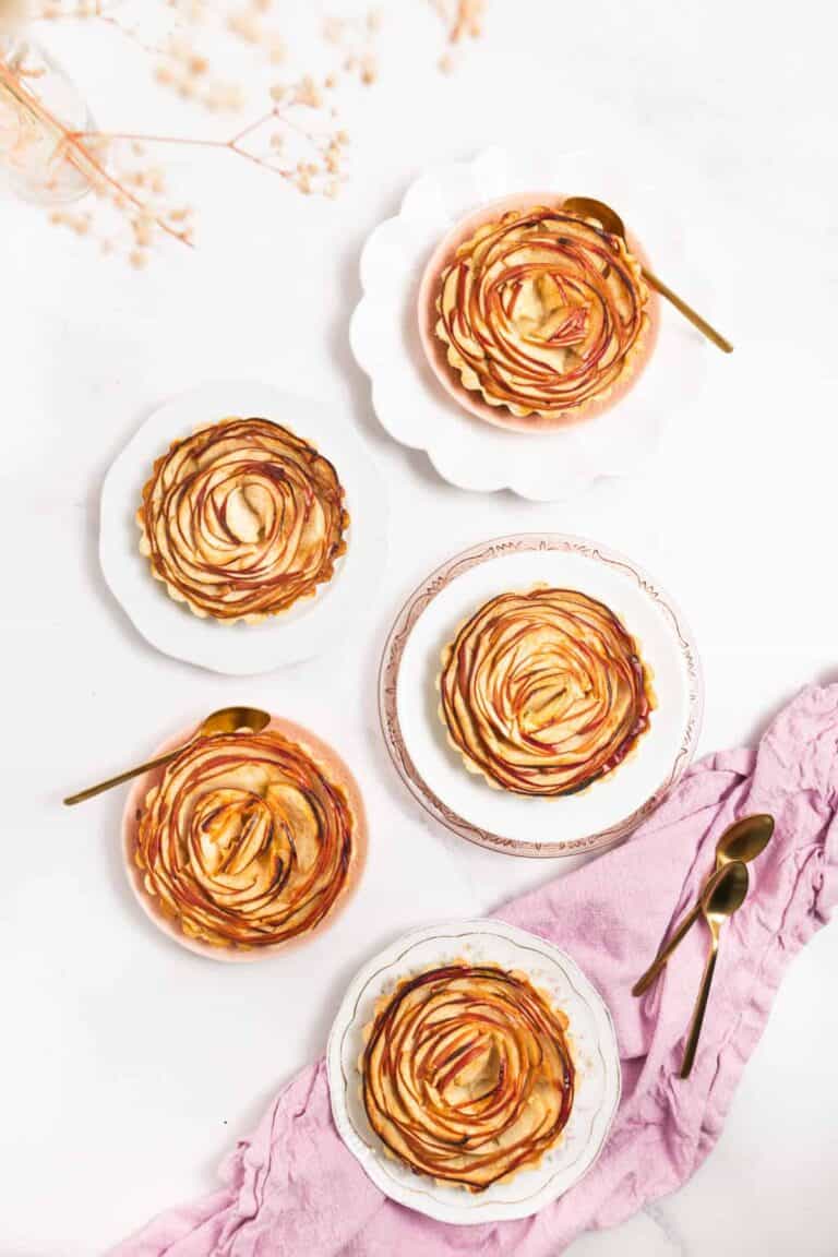 Five elegantly arranged apple tarts on scalloped plates with gold spoons, resting on a light marble surface. A pink cloth lies beside them with additional spoons, creating a cozy and inviting scene.