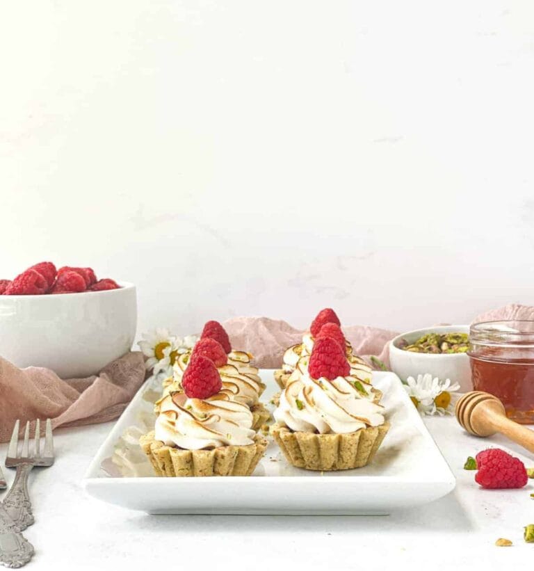 Mini tartlets topped with swirls of white cream and fresh raspberries on a white rectangular plate. A bowl of raspberries, a jar of honey with a dipper, and a dish of nuts are in the background. Soft pink cloth and two forks are nearby.