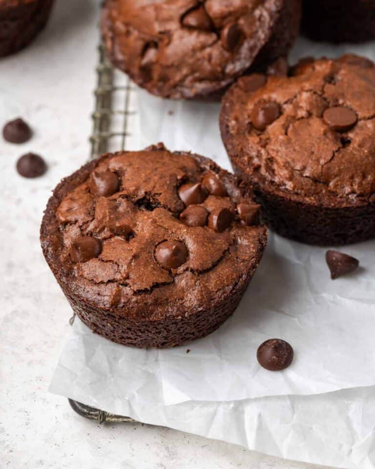 Two chocolate muffins with chocolate chips sit on crinkled parchment paper atop a wire rack. More chocolate chips are scattered around. The muffins have a cracked, crumbly texture.