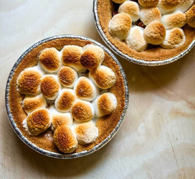 Two small pies in foil tins topped with toasted marshmallows on a light marble surface.