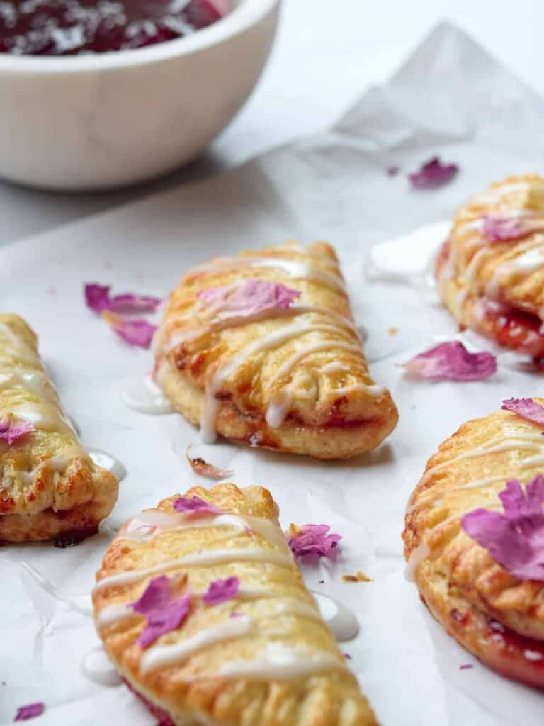 Close-up of glazed pastries with pink flower petals on top, arranged on parchment paper. A bowl of raspberry jam is blurred in the background. The pastries have a golden-brown crust with light icing drizzles.