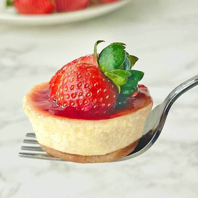 A fork holds a small cheesecake topped with glossy strawberry sauce and a fresh strawberry. The background is a soft focus, white marble surface.