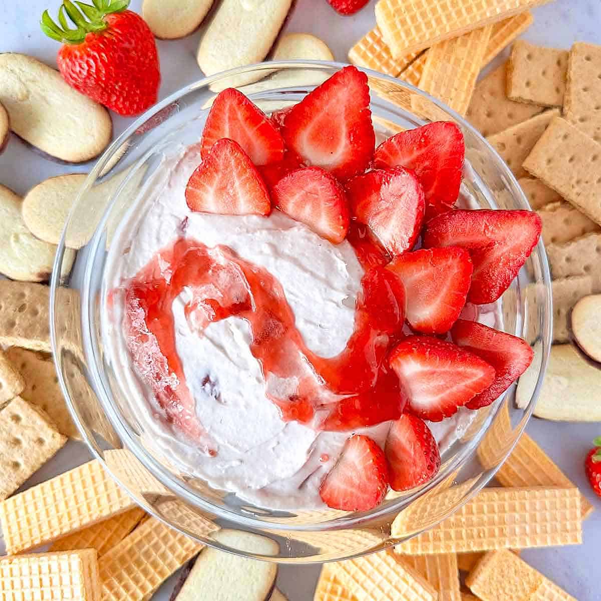 A glass bowl of strawberry cheesecake dip with fresh strawberries and cookies for dipping.