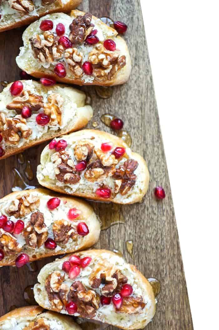 Six goat cheese crostini topped with pomegranate seeds, on a cutting board.