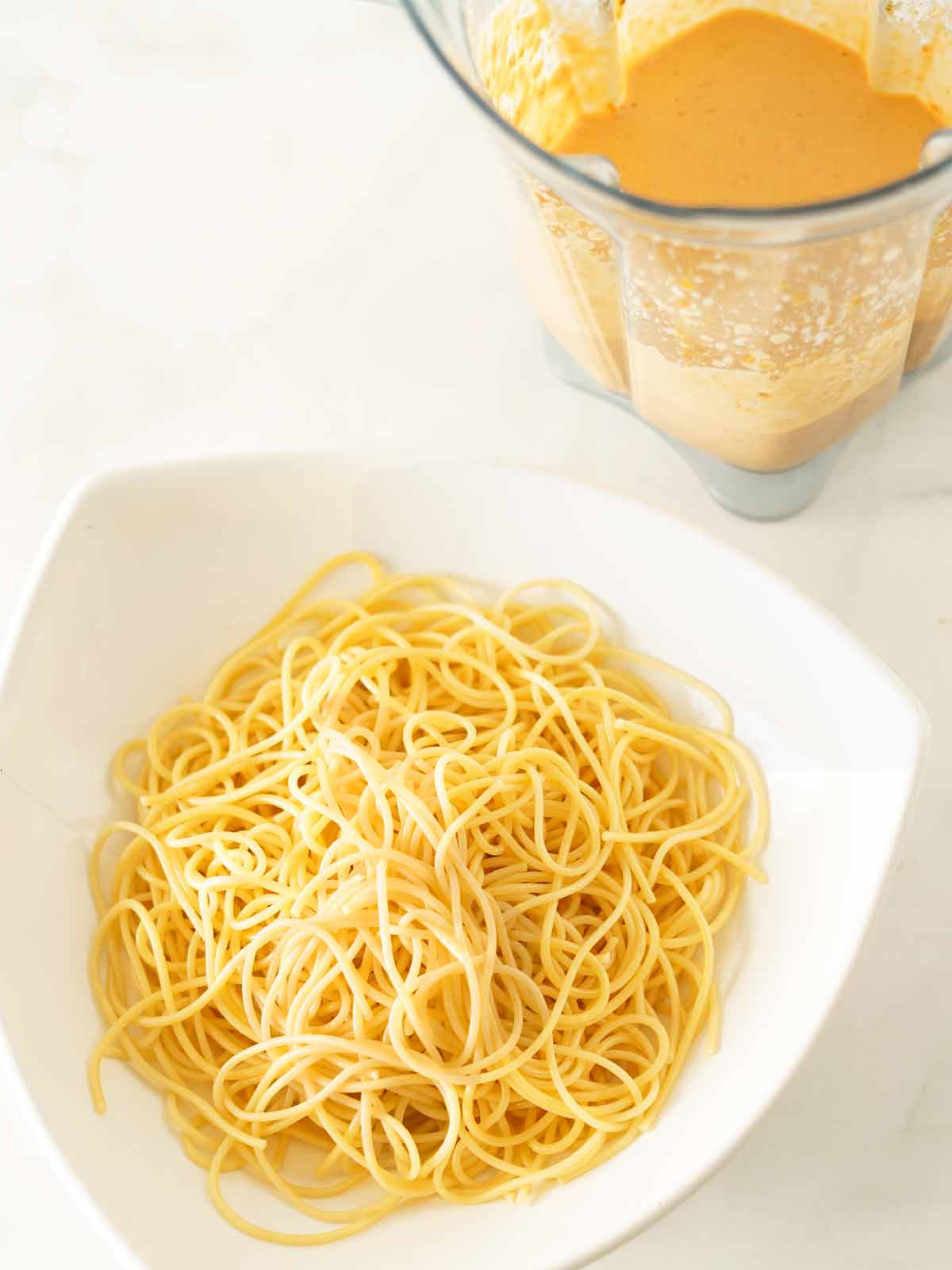 Burrata sauce with tomatoes in the blender and a white bowl of hot pasta noodles.