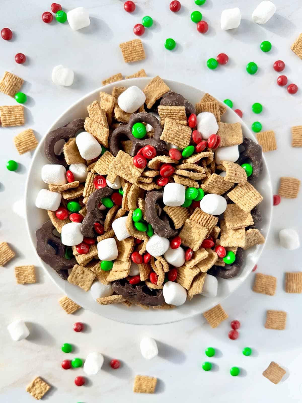 A bowl of S'mores snack mix surrounded by marshmallows, golden graham cereal, and M & Ms candies.