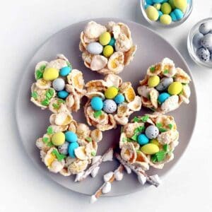 Six birds nest cookies on a gray serving dish, and two bowls of chocolate mini eggs.