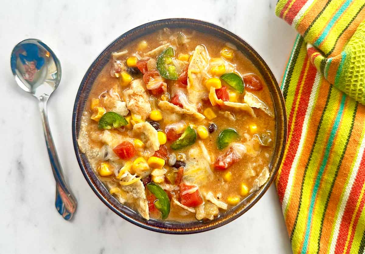 A soup spoon, a green dishtowel, and a brown bowl of creamy chicken tortilla soup.
