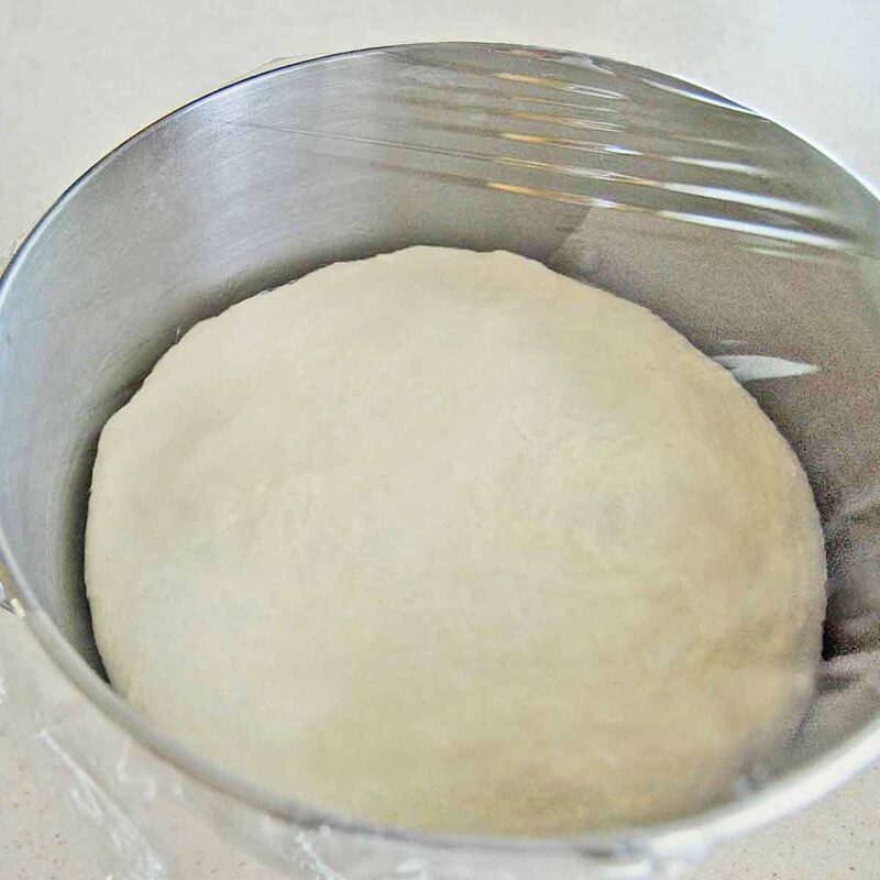 Homemade pizza dough in a stainless steel mixing bowl covered with plastic wrap and rising.