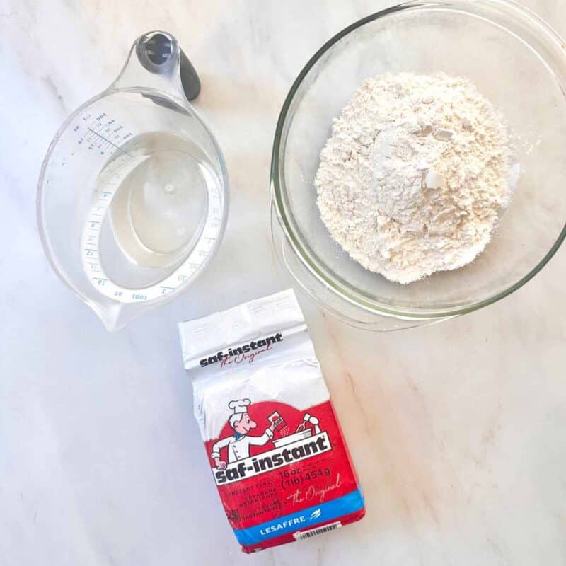 A measuring cup with water, a bowl with bread flour, and a bag of instant yeast.