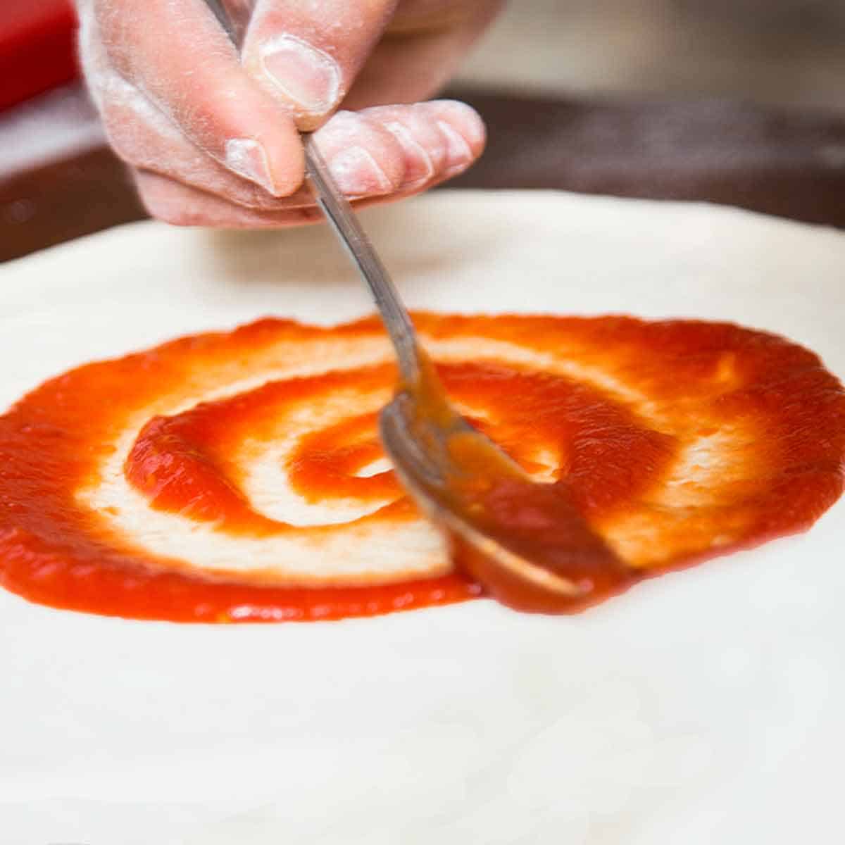 Using a spoon to spread pizza sauce on NY style pizza dough.