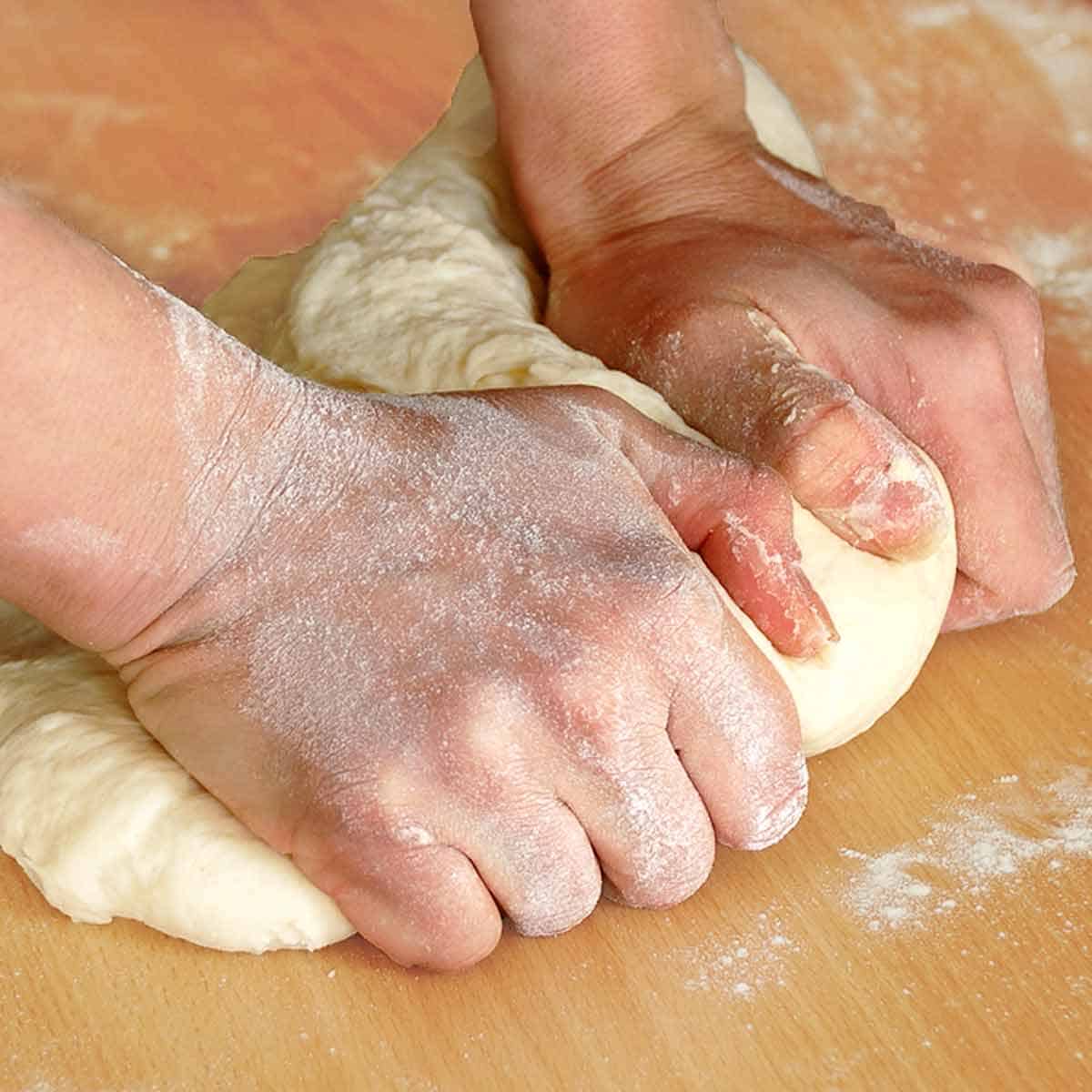 Two hands kneading pizza dough on a wood cutting board.
