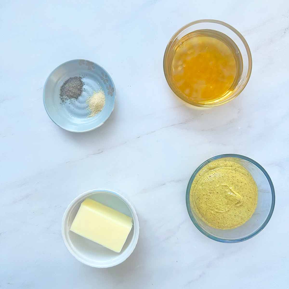 Four bowls containing honey, Dijon mustard, unsalted butter, ground black pepper and garlic powder.